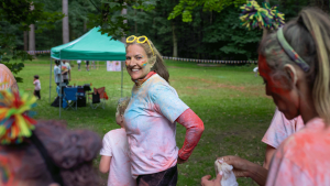 Medium shot of a grandma and her grandson standing together after an LGBTQ+ colour run in a woodland area in Gateshead. They are standing with another family, and they are all covered in colourful paint. The grandma is looking at the camera and smiling while wearing colourful sunglasses on her head.